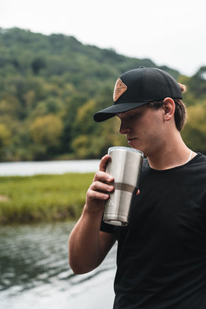 A young man wearing a black Kentzo Koffee hat and t-shirt holds a stainless steel tumbler close to his face, standing by a peaceful riverbank with lush, green trees and hills in the background.